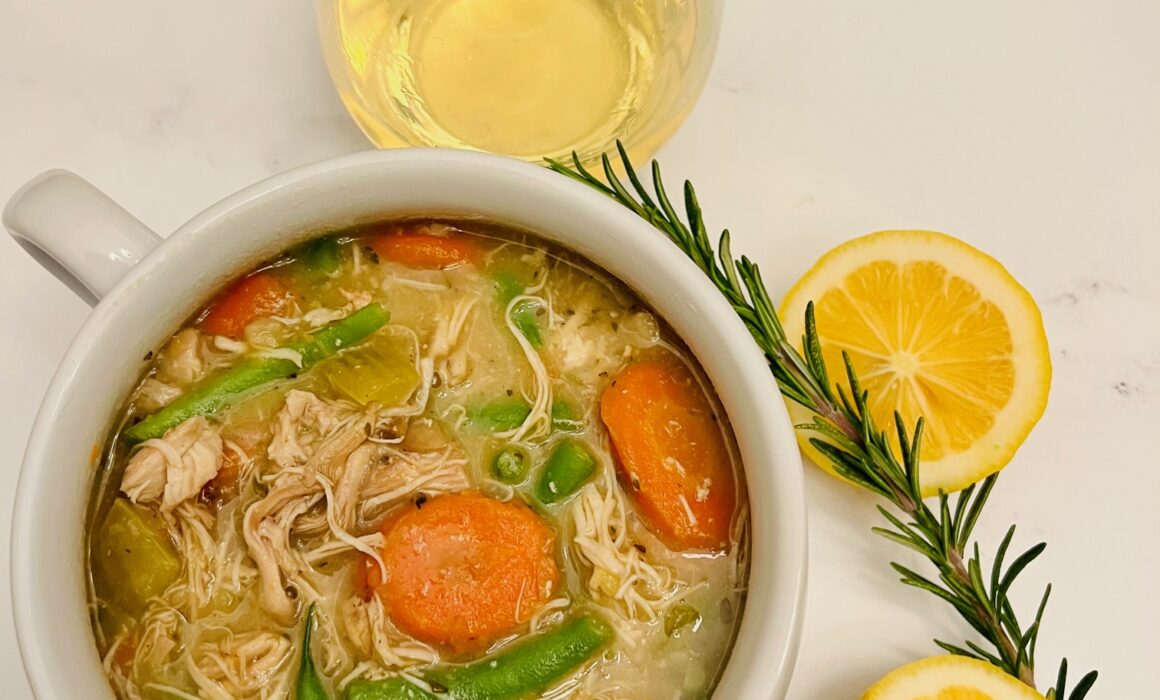 Slow-cooker Chicken Stew in a bowl with a glass of white wine. On the table are two slices of lemon and a sprig of rosemary