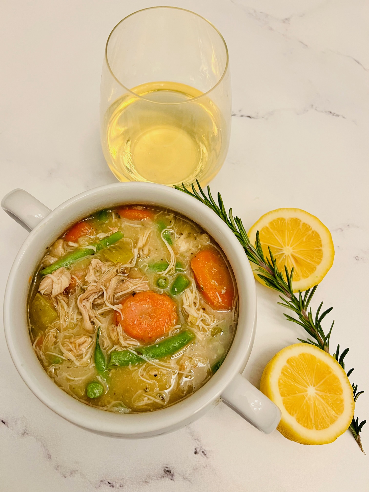 Slow-cooker Chicken Stew in a bowl with a glass of white wine. On the table are two slices of lemon and a sprig of rosemary