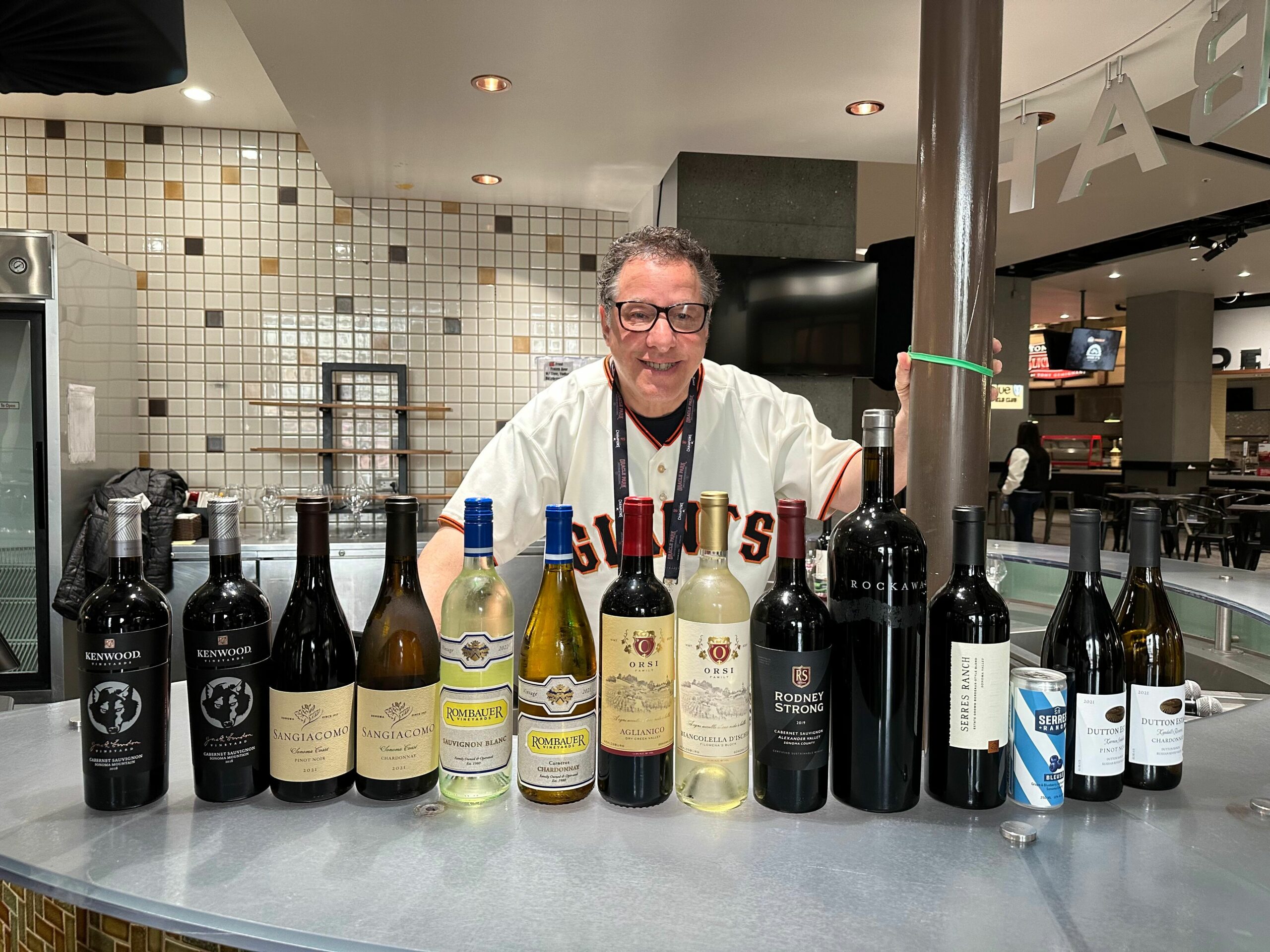 Man standing behind a bar, with 13 bottles of wine in front of him