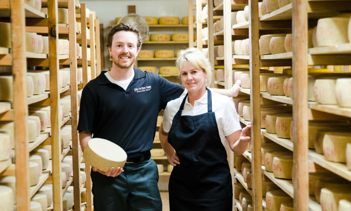 Filled cheese cellar and two people holding a wheel of cheese