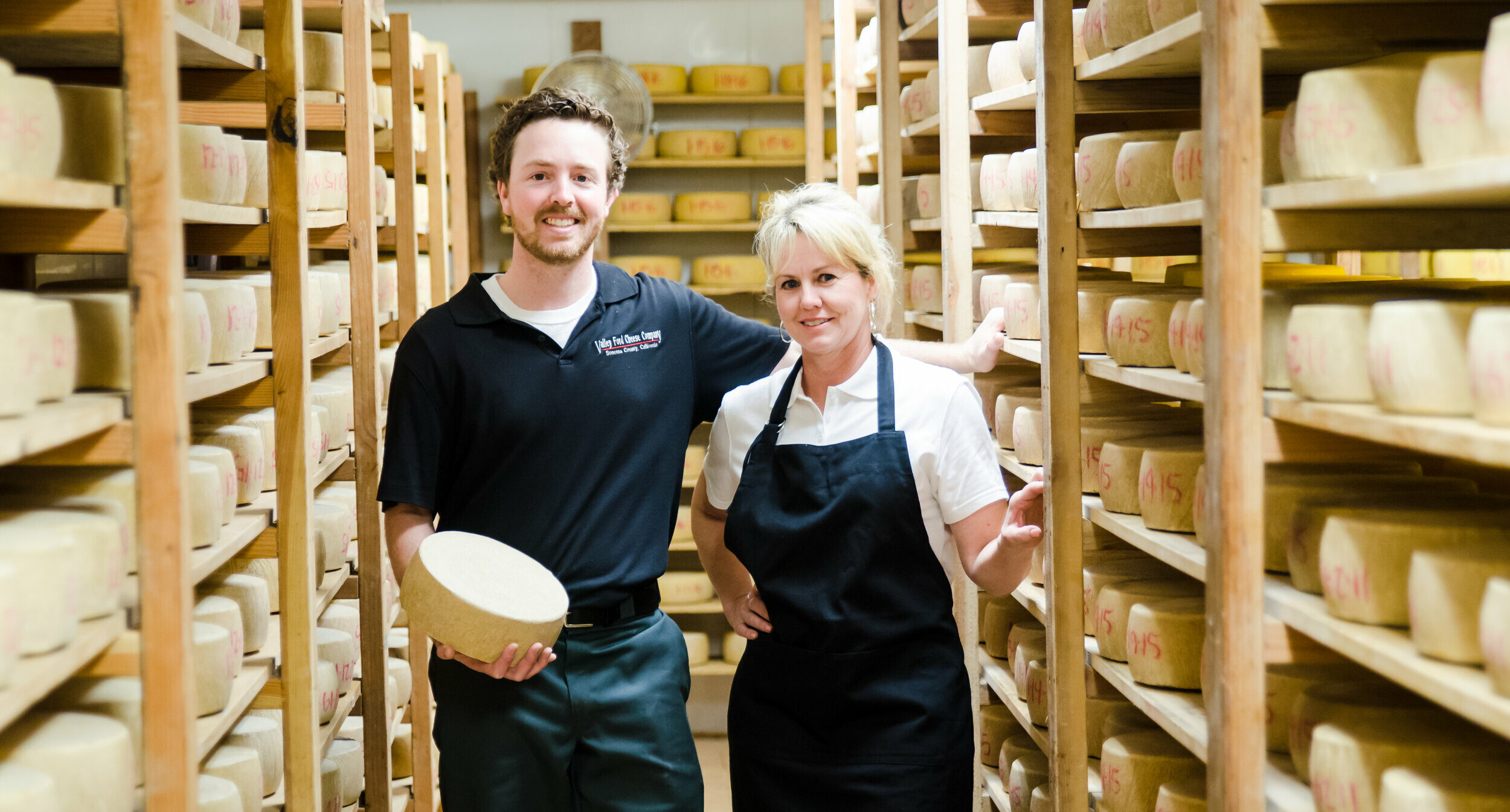 Filled cheese cellar and two people holding a wheel of cheese