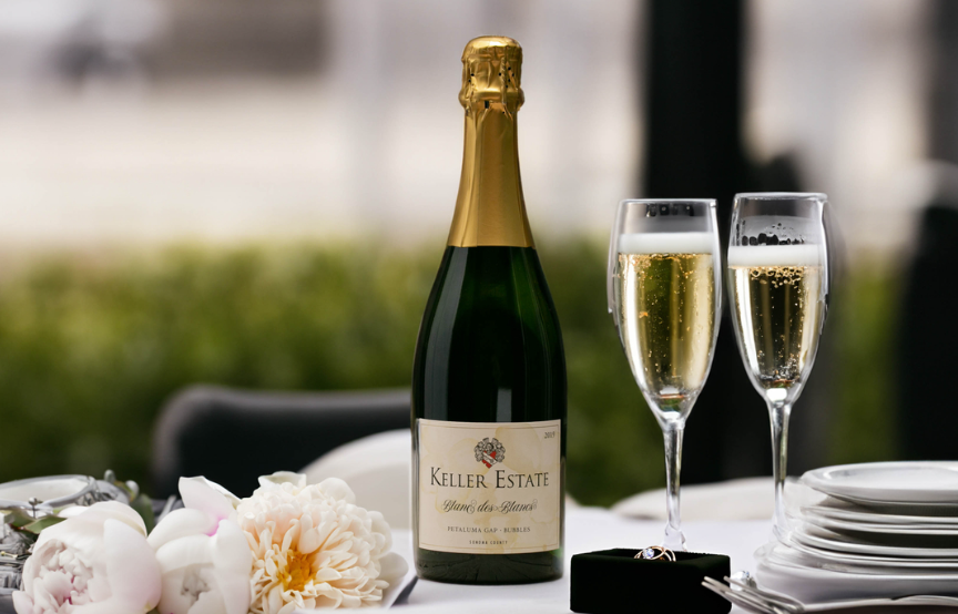 Bottle of Keller Estate Brut beside two filled champagne flutes and a stack of white plates